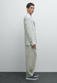 Completo in lino grigio chiaro con giacca a un bottone e pantaloni a gamba larga. Abbinato a scarpe slip-on grigio scuro. Design e texture minimalisti.