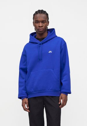 Junger Mann mit geflochtenem Haar, der einen hellblauen Nike SB Hoodie und schwarze Hose trägt und vor einem weißen einfarbigen Hintergrund steht.