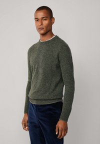 Dunkelgrüner gestrickter Pullover mit Rundhalsausschnitt, gerippten Bündchen und Saum, kombiniert mit navyblauen Cordhosen. Glatte Textur mit einem subtilen Muster.