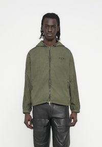 Χακί φούτερ με φερμουάρ και κουκούλα, με υφή στις πλαϊνές ρίγες και διακοσμητικό λογότυπο. Φορεμένο με μαύρο δερμάτινο παντελόνι cargo.