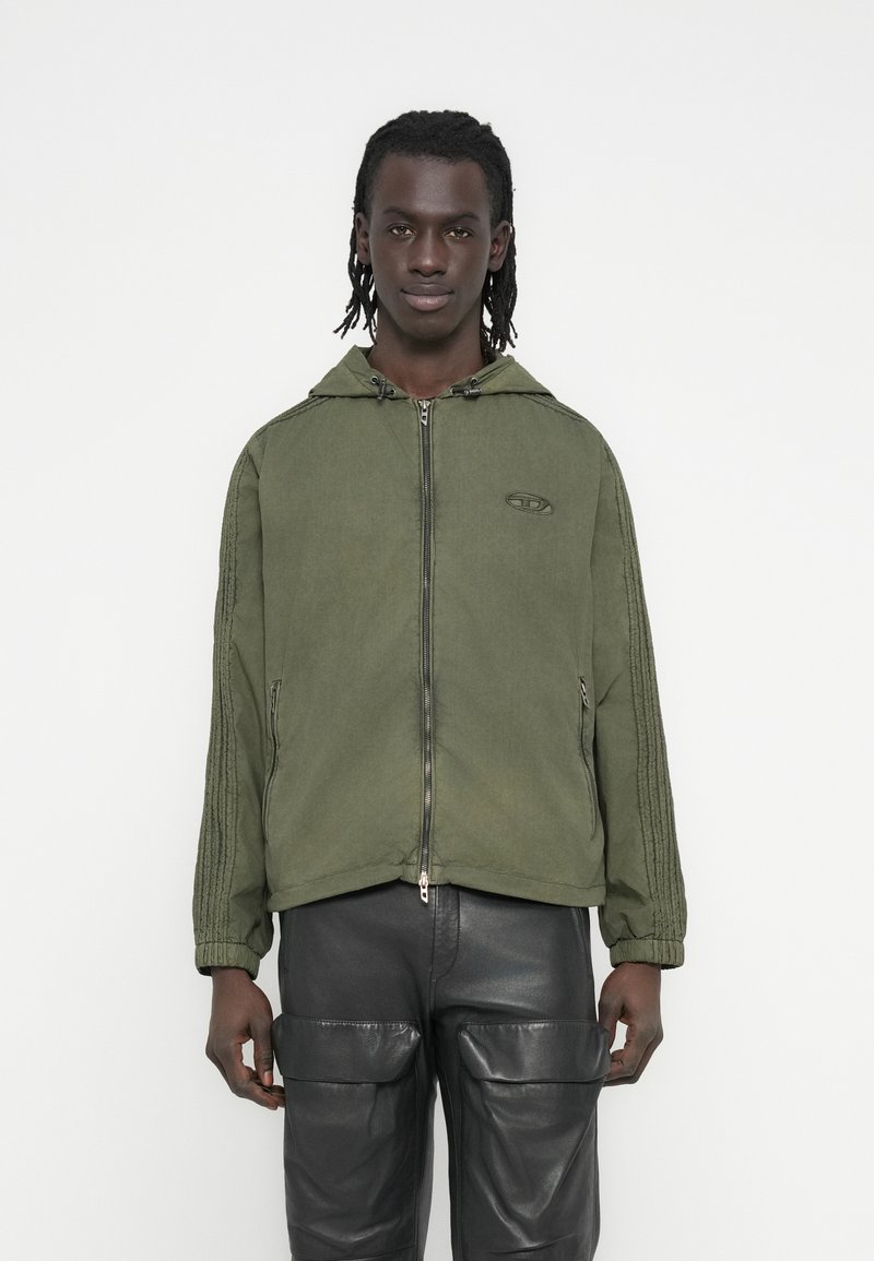 Χακί φούτερ με φερμουάρ και κουκούλα, με υφή στις πλαϊνές ρίγες και διακοσμητικό λογότυπο. Φορεμένο με μαύρο δερμάτινο παντελόνι cargo.