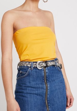 Femme portant un haut jaune sans bretelles, une jupe en jean bleue taille haute avec une fermeture éclair centrale, et une ceinture tressée argentée avec une boucle décorative.