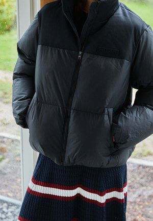 Giacca nera imbottita con zipper frontale, caratterizzata da una parte superiore uniforme. La indossatrice la abbina a una gonna a coste con strisce orizzontali.