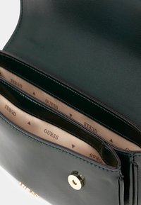 Interior de bolso de mano de cuero verde con múltiples compartimentos, con un suave forro beige impreso con "GUESS" y un cierre a presión dorado.