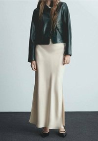 Černá kožená zkrácená bunda, béžová saténová maxi sukně s postranním rozparkem a černé ploché boty. Viditelný perlový náhrdelník zdůrazňuje vrstvené textury.