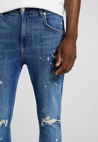 Blå denimjeans med slitna detaljer, inklusive hål och vita färgstänk. Har en klassisk femfickdesign och dragkedja.