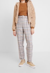 Beige Kunstfelljacke, brauner glitzernder Pullover, karierte Hose mit beige-, grau- und gelbtönen Akzenten und beige Sneakern.