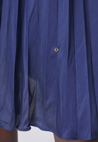 Falda azul plisada hecha de tela ligera con un brillo. Presenta un pequeño logotipo dorado cerca del dobladillo. Textura suave y cinturilla ajustable.