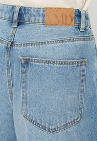 Helle blaue Jeans aus Denim mit kontrastierendem gelbem Stitching, ausgestattet mit einer Gesäßtasche und einem braunen Lederpatch, das mit "VMD" geprägt ist.