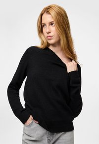 Jauna moteris su ilgais šviesiais plaukais dėvi juodą megztinį ir šviesiai pilkas džinsus, viena ranka laiko megztinio apykaklę, žiūri į kairę.