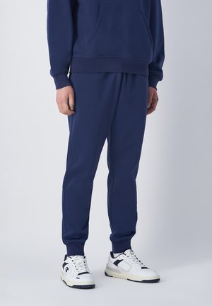 Sweatshirt navy avec une poche frontale, des poignets élastiques et un pantalon assorti. Des sneakers blanches avec des accents noirs complètent la tenue.