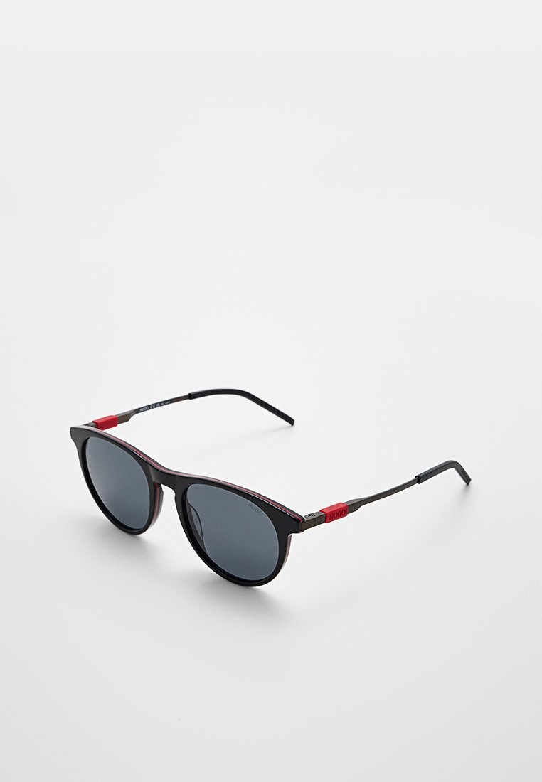 Schwarze Sonnenbrille mit grauen Gläsern. Katzenaugenform, dünne schwarze Bügel mit roten Akzenten. Leichtes Material, minimalistisches Design.