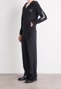 Felpa nera con zip e fodera arancione, con logo "BOSS" e design a onda sonora sulla manica. Indossata con pantaloni a pieghe neri e scarpe.