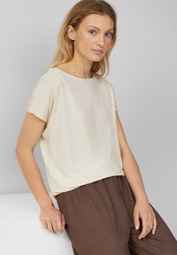 Vrouw met lichtblond haar die een crèmekleurig shirt met korte mouwen en bruin gestructureerde broek draagt, leunend op een witte oppervlakte tegen een grijze achtergrond.