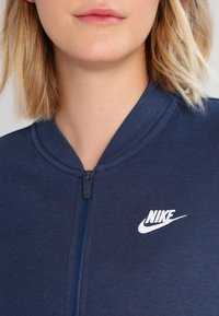 Mörkblå zip-up jacka med hög krage, tillverkad av ett strukturerat tyg. Har en vit Nike-logotyp på vänster bröstområde.