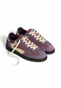 Un par de zapatillas moradas, beige y burdeos con cordones blancos, suelas negras y el nombre de la marca en la lengüeta, con un cordón desatado sobre un fondo blanco.
