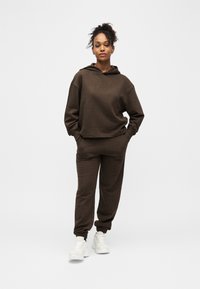 Γυναίκα που στέκεται με τα χέρια στις τσέπες, φοράει καφέ φούτερ με κουκούλα και ταιριαστό παντελόνι τζόγκινγκ, καθώς και λευκά αθλητικά παπούτσια, σε απλό λευκό φόντο.