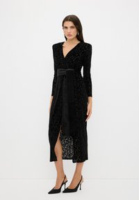 Schwarzes Samtwickelkleid mit langen Ärmeln, tiefem V-Ausschnitt, einem satinierten Gürtel und einem Oberschenkel-hohen Schlitz mit aufwändigen strukturierten Mustern.