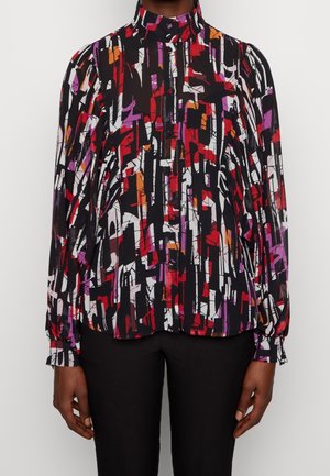 Bluzka z długim rękawem i wysoką szyją z abstrakcyjnym geometrycznym nadrukiem w kolorach czarnym, białym, czerwonym, fioletowym i pomarańczowym, noszona z czarnymi spodniami.