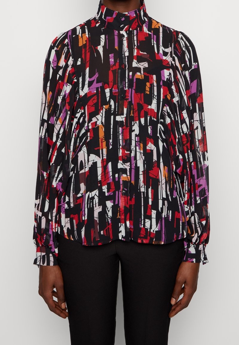 Blouse à manches longues et col haut avec imprimé géométrique abstrait en noir, blanc, rouge, violet et orange, portée avec un pantalon noir.