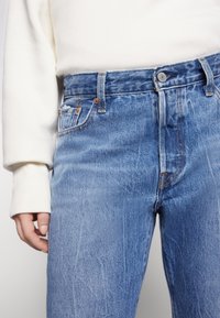 Jeans de mezclilla azul deslavado con un corte clásico, que presentan un cierre frontal con botón, cinco bolsillos y acentos de costura en naranja.