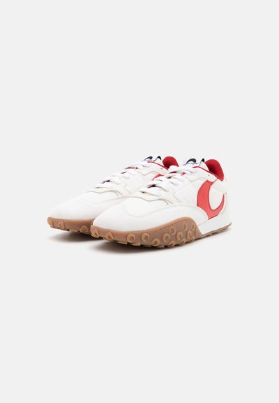 White sneakers with red accents, textured fabric, and a gum rubber sole featuring circular patterns. Laces are white; logo on tongue is black.