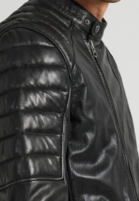 Svart lädermotorcykeljacka med ett kviltat axeldesign, hög krage med tryckknappstängning och en framträdande dragkedja framtill.