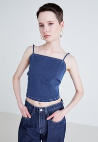 Denim crop top s tenkými ramienkami, s obdĺžnikovým výstrihom a surovým lemom. Model ho kombinuje s tmavomodrými nohavicami s vysokým pásom.