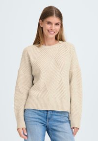 Beige gestrickter Pullover mit gerippter Textur und Rautenmuster. Verfügt über einen runden Halsausschnitt und lange Ärmel, kombiniert mit blauen Jeans.