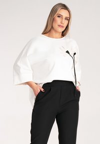 Biała, oversize'owa bluzka z okrągłym dekoltem, z kwiatowym akcentem i czarnymi detalami; w połączeniu z dopasowanymi czarnymi spodniami.