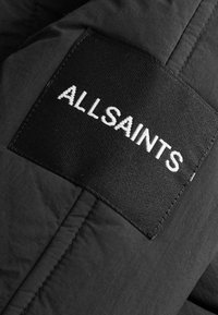 Černá látková nášivka s bílým textem "ALLSAINTS", s texturovaným povrchem a šitými okraji, připevněná k části svrchního oděvu.