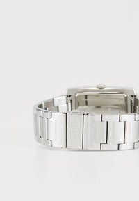 Montre-bracelet en métal argenté avec un boîtier rectangulaire, un bracelet à maillons et une surface texturée. Le nom de la marque est gravé sur le fermoir.