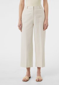 Beżowe spodnie o szerokich nogawkach z wysokim stanem, z czystym przodem i teksturą materiału. Połączone z beżowymi plecionymi sandałami.