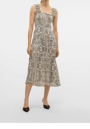 Femme portant une robe midi beige sans manches avec des motifs géométriques abstraits noirs et des chaussures noires pointues sur un fond uni.
