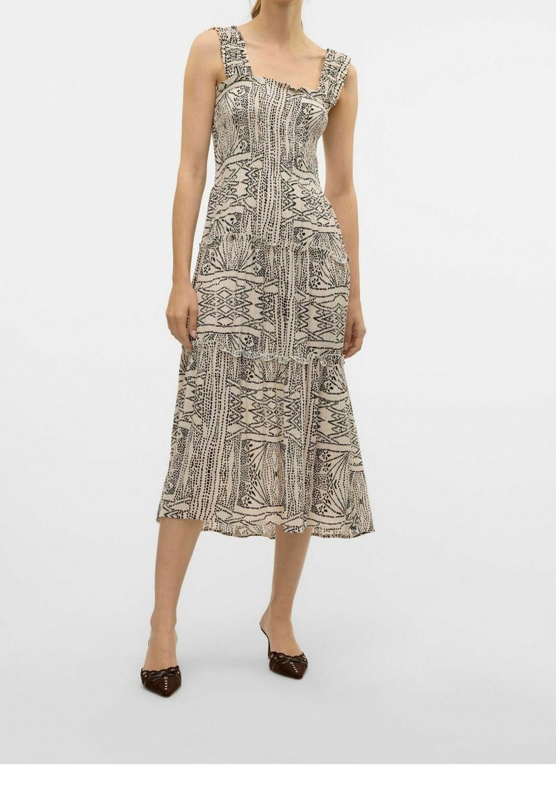 Femme portant une robe midi beige sans manches avec des motifs géométriques abstraits noirs et des chaussures noires pointues sur un fond uni.