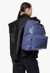 Zaino in tessuto sintetico blu con design arrotondato, tasca frontale con zip e spallacci regolabili, indossato sopra una giacca di pelle nera.