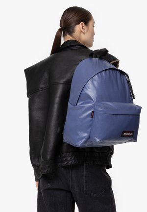 Mochila de tela sintética azul con un diseño redondeado, bolsillo frontal con cremallera y correas ajustables, llevada sobre una chaqueta de cuero negra.