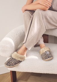 Leopardimustriga sussid beežika kunstkarusnaha mansetiga, pehme tekstuuriga pealispind ja helepruun kummist tald, koos halli kodurõivastusega pehmel tugitoolil.