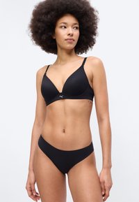 Γυναίκα με αφροκάν μαλλιά που φοράει μαύρο σουτιέν και σλιπ που ταιριάζουν, σε λευκό φόντο, κοιτάζοντας ελαφρώς στο πλάι.