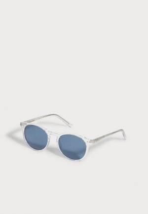 Lunettes de soleil à monture transparente avec des verres ronds bleus reposant sur une surface blanche, légèrement inclinées vers la droite.