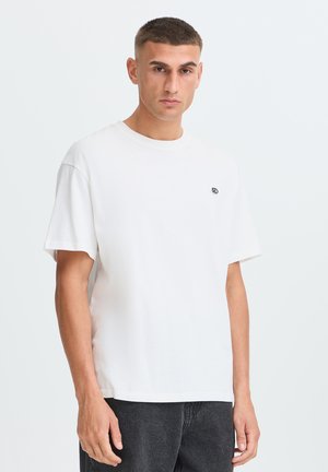 Joven con el pelo corto que lleva una camiseta blanca lisa con un pequeño logo en el pecho y pantalones oscuros sobre un fondo liso.