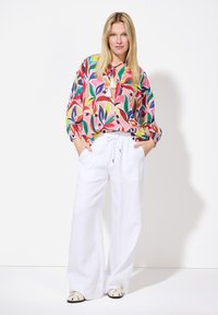 Bunter Blumenbluse mit langen Ärmeln, kombiniert mit lockeren weißen Leinenhosen und flachen beigen Sandalen. Die Bluse zeichnet sich durch lebendige Blattmuster aus.