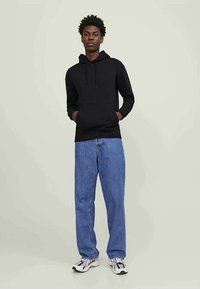Giovane uomo che indossa un hoodie nero, jeans blu larghi e sneakers bianche, in piedi con le mani nelle tasche contro uno sfondo neutro.