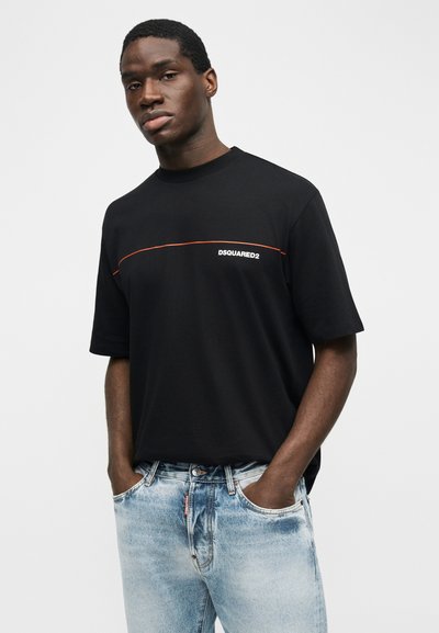Νέος άνδρας στέκεται με τα χέρια στις τσέπες, φορώντας ένα μαύρο t-shirt DSQUARED2 και ανοιχτόχρωμο μπλε τζιν, μπροστά σε απλό φόντο.
