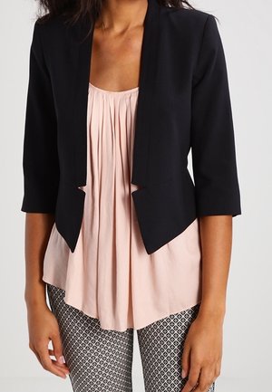 Frau trägt einen schwarzen, bauchfreien Blazer über einer blassrosa plissierten Bluse mit gemusterten schwarz-weißen Hosen.