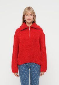 Dieprood gestructureerd sweater met een hoge kraag, een halve ritssluiting aan de voorkant en verlaagde schoudernaad. Draagt over blauwe jeans met een diamanten patroon.