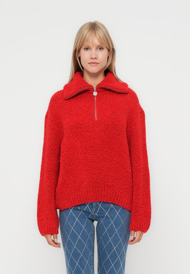 Dieprood gestructureerd sweater met een hoge kraag, een halve ritssluiting aan de voorkant en verlaagde schoudernaad. Draagt over blauwe jeans met een diamanten patroon.