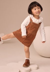 Vestito in velluto a coste marrone con spalline con bottoni, camicia bianca con volant sotto, stivaletti marroni con lacci, in piedi su una piattaforma a forma di fiore.