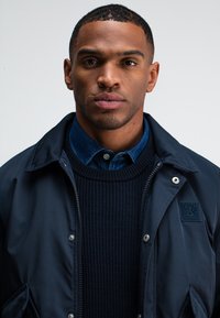 Marineblaue Zip-Jacke mit Druckknöpfen, darunter ein strukturiertes schwarzes Sweatshirt und ein Jeanshemd mit Kragen. Subtiles Logo auf der Jacke.