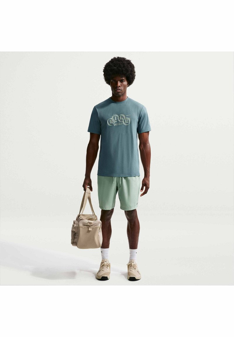 Homme avec coiffure afro portant un t-shirt graphique bleu, un short vert clair, des baskets beiges, des chaussettes blanches, tenant un sac de sport beige, debout sur fond blanc.